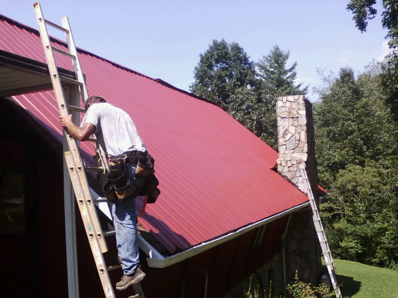 Roofer on a ladder beside a red metal roof and stone chimney — Soffit & Fascia Repair by Roofing Brothers in Pe Ell
